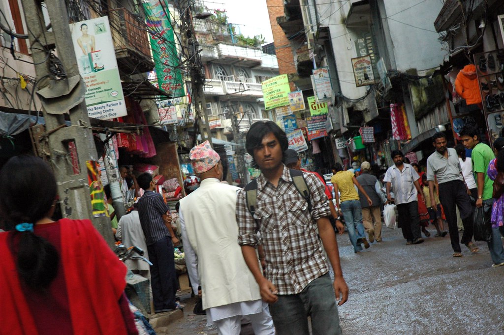 A man walks down the street wearing a plaid shirt and a backpack, there is a story in this photo, people, signs, scary electrical wiring, shops, clothes,…