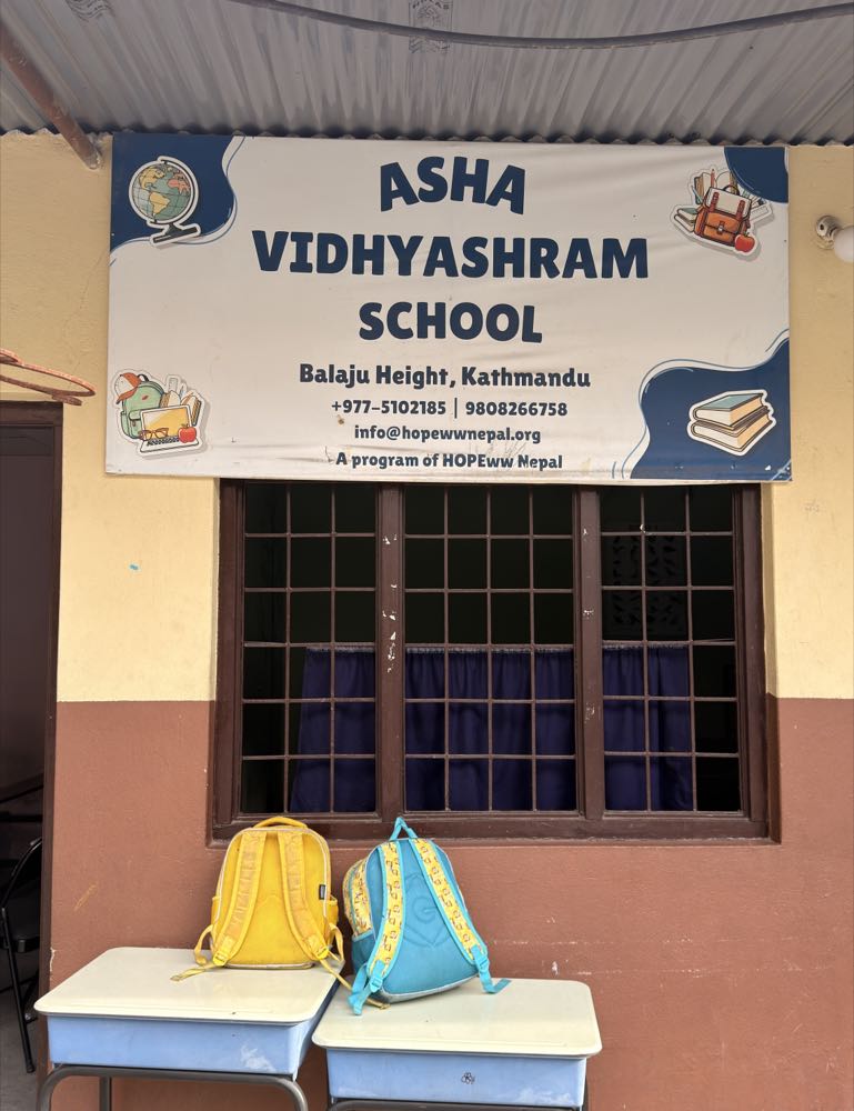 The Asha Vidhyashram School sign at Balaju Height, Kathmandu, with two children's backpacks resting on desks beneath it.