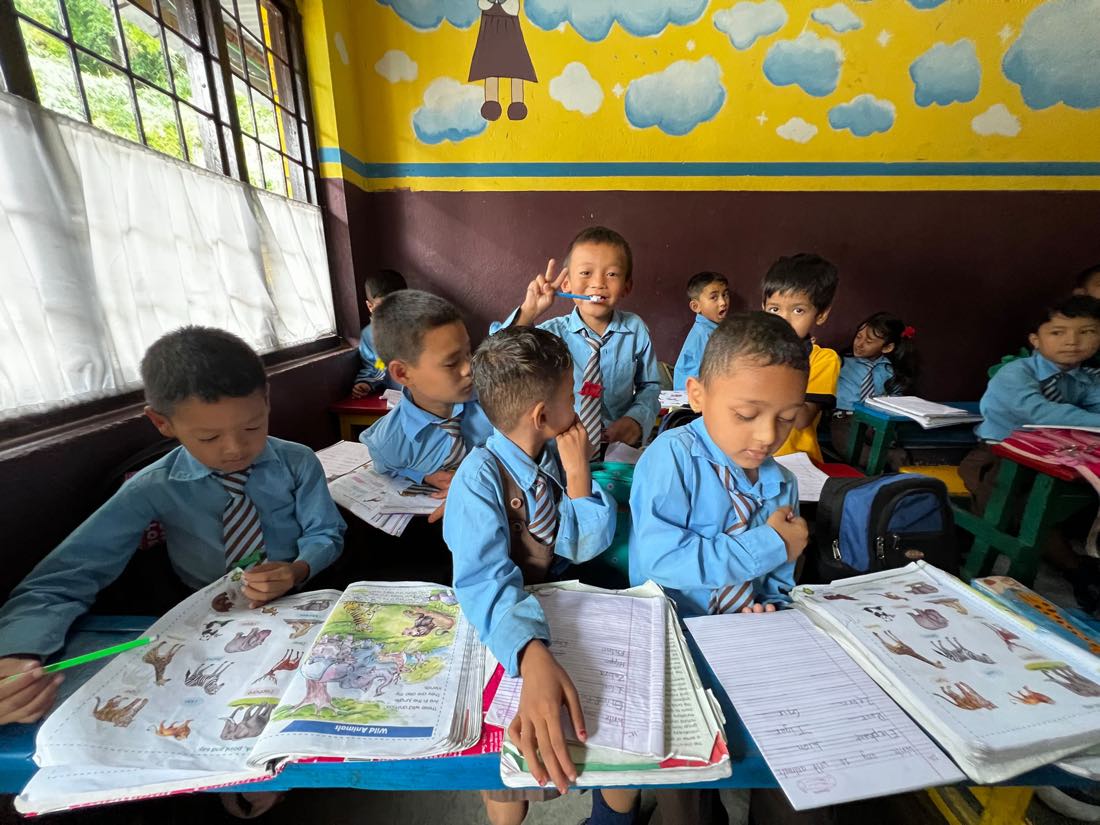 Students in blue uniforms at their desks with workbooks open to a page of animals, one boy holding up a toothbrush, painted clouds along the classroom wall.