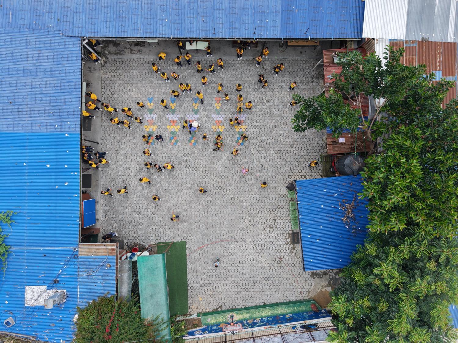 Around a hundred children in yellow uniforms gathered across the Asha Vidhyashram School courtyard, seen from directly above.