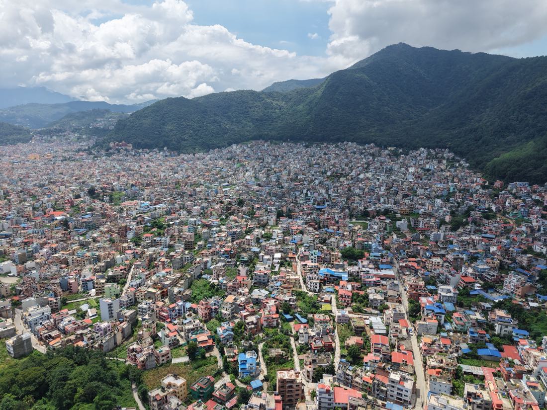 The Kathmandu valley from the air, dense housing running up to a forested ridge under heavy cloud.