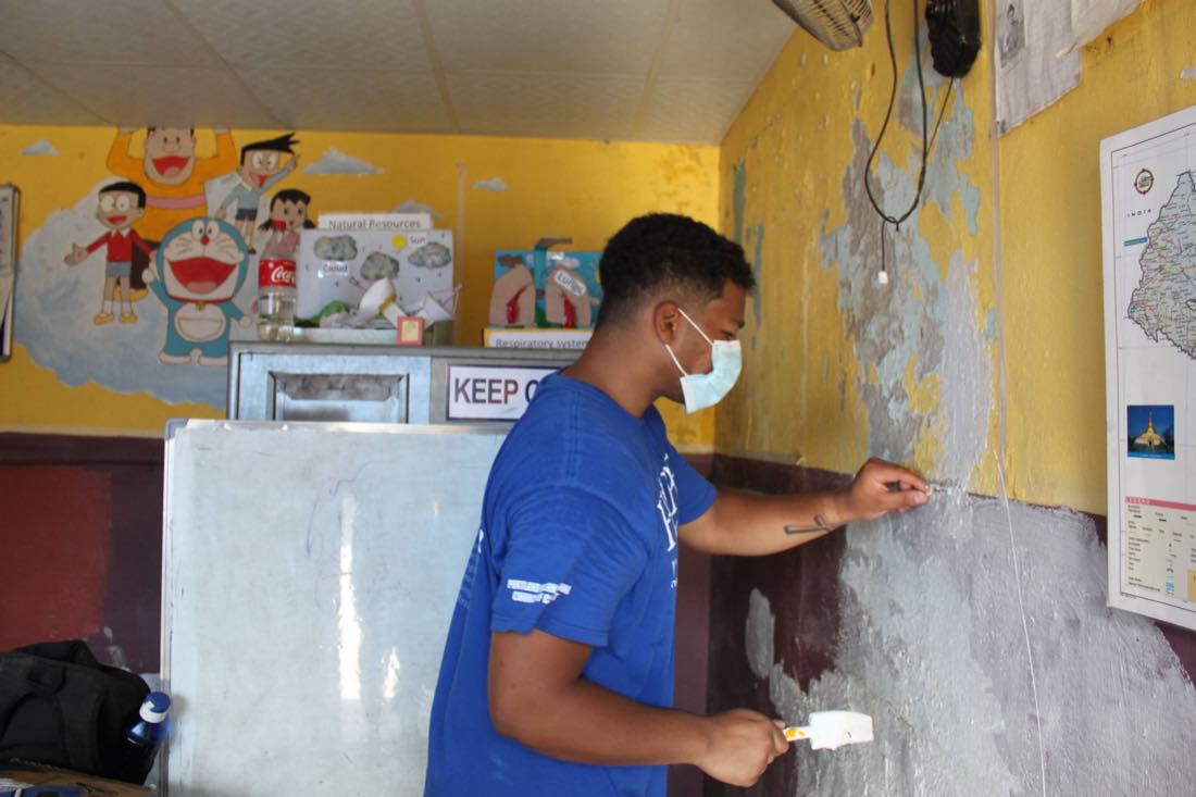 Shawn Reddy patching and repainting a classroom wall at the Asha school, teaching charts and a map still on the wall beside him.