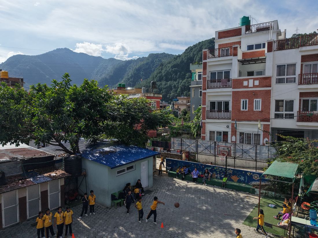 The school yard set among houses on the Kathmandu hillside, forested ridges rising beyond the rooftops.