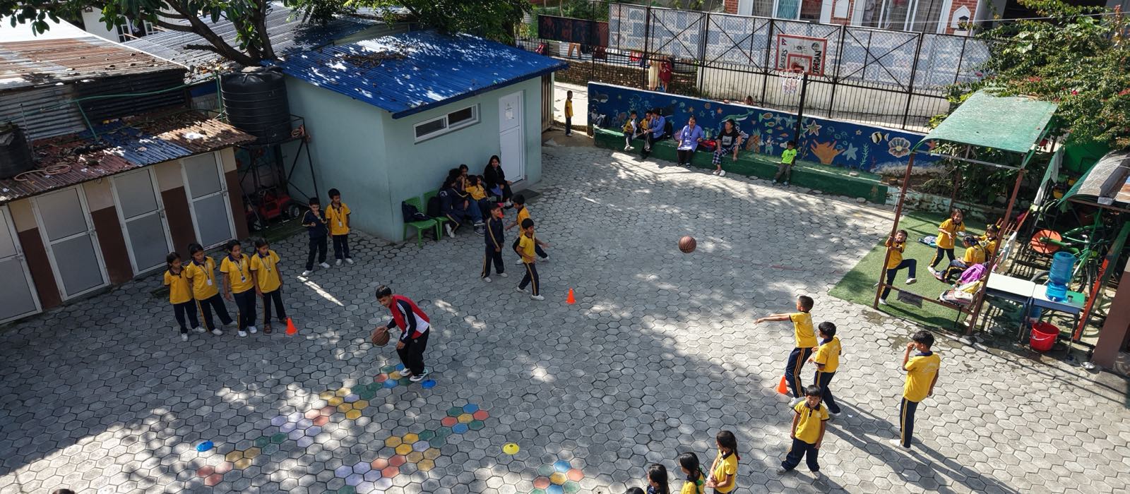 Children in yellow uniforms playing across the Asha Vidhyashram School yard, with Kathmandu houses and hillside rising behind the school.