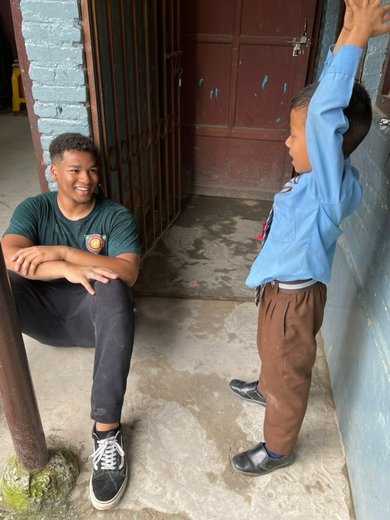 Shawn Reddy sitting on the ground in a school doorway, smiling up at a young student in a blue uniform who is reaching a hand above his head.