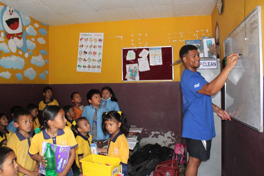 Shawn Reddy at the whiteboard in an Asha classroom, a group of students in yellow and blue uniforms watching from the floor.